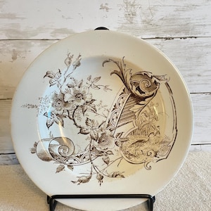 May include: A round ceramic plate featuring a dark brown transferware pattern on a cream background. The design includes detailed illustrations of wild roses, scrolling acanthus leaf motifs, and a small scene of a bird in a landscape.
