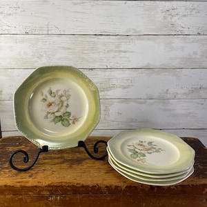 May include: A set of vintage octagonal plates with a floral rose design. The plates have a pale green border and a cream-colored center. One plate is displayed on a black metal stand, and a stack of plates is on a wooden surface.