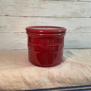 May include: A red ceramic jar with a matching lid. The jar has a cylindrical shape with a textured, basket-weave design around the body. The jar is sitting on a white and blue striped cloth.