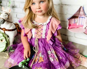 May include: A young girl dressed as Rapunzel, wearing a purple dress with floral embroidery and a flower crown. She is holding a frying pan with a green chameleon on it and a sun design on the bottom.