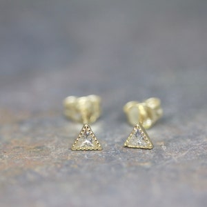 May include: A pair of gold-colored triangle stud earrings. Each earring features a clear, triangular-cut stone set within a beaded frame. The earrings are displayed on a textured, gray surface.