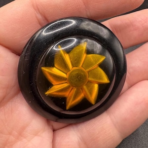 May include: A circular brooch featuring a smooth, glossy black outer rim and a central, raised golden-yellow star-shaped element with eight pointed petals. The design is set against a dark, reflective background, creating a high-contrast appearance.