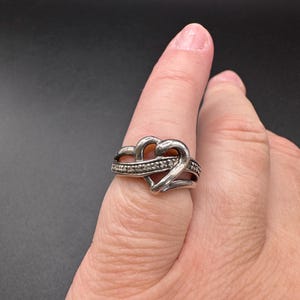 May include: A silver ring with a heart design. The heart is made of two intertwined bands, and the ring is set with small, clear stones.