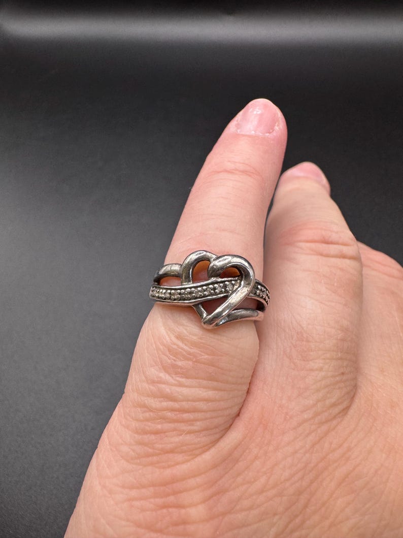May include: A silver ring with a heart design. The heart is made of two intertwined bands, and the ring is set with small, clear stones.