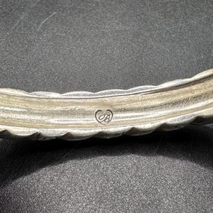 Puede incluir: Primer plano de una pulsera plateada con un dise&ntilde;o texturizado y festoneado. La pulsera presenta un emblema en forma de coraz&oacute;n con las iniciales "JB". El metal tiene un aspecto ligeramente deslustrado.