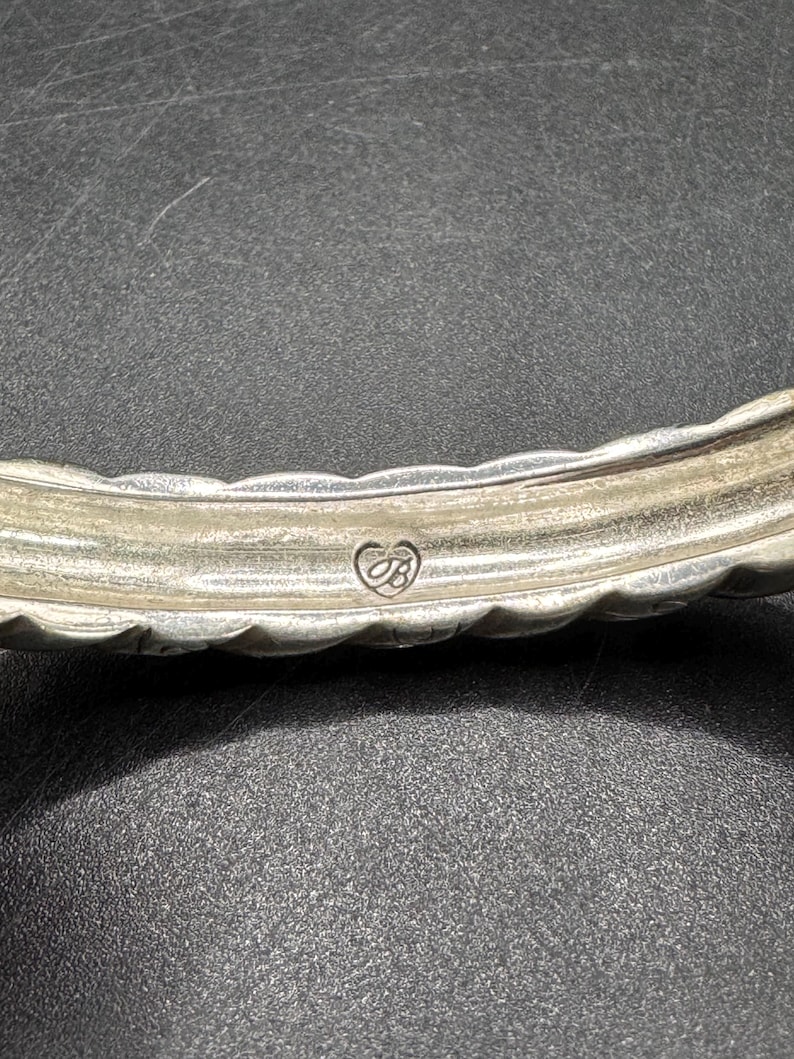 Puede incluir: Primer plano de una pulsera plateada con un dise&ntilde;o texturizado y festoneado. La pulsera presenta un emblema en forma de coraz&oacute;n con las iniciales "JB". El metal tiene un aspecto ligeramente deslustrado.