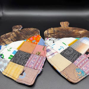 May include: Two acorn-shaped fabric potholders featuring a patchwork design of various patterned squares in colors including white, blue, orange, pink, and brown. The top section of each potholder is made of a brown fabric with a textured pattern.