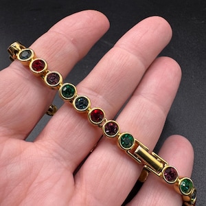 May include: A gold-tone metal bracelet featuring a series of round, bezel-set faceted stones in alternating colors of red, green, purple, and dark blue. The bracelet includes a secure metal clasp mechanism and a polished finish throughout the links.