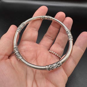 May include: A silver bangle bracelet featuring decorative etched patterns. The bracelet has a smooth, rounded design with detailed ornamental sections. The bracelet is shown in a hand.
