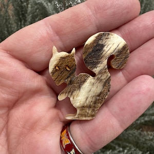 May include: A wooden squirrel figurine with a curved tail. The squirrel is carved from a light-colored wood with brown marbling. The figurine has small, dark eyes and is held in a hand.