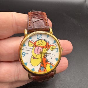May include: A gold-tone analog wristwatch featuring a white dial with a graphic illustration of Tigger. The watch has blue numeral markers at 12, 3, 6, and 9, with blue triangle indices between them. It includes a brown textured leather strap.
