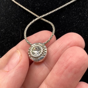 May include: A silver pendant necklace with a round, textured pendant. The pendant features a central clear stone surrounded by a sunburst design. The chain is a delicate, woven style.