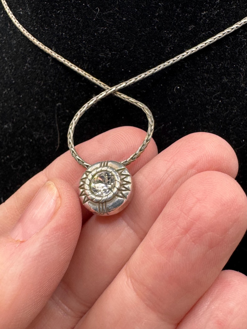 May include: A silver pendant necklace with a round, textured pendant. The pendant features a central clear stone surrounded by a sunburst design. The chain is a delicate, woven style.