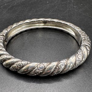 Puede incluir: Un brazalete plateado con un dise&ntilde;o de cuerda trenzada y adornos ornamentados. El brazalete tiene una marca en forma de coraz&oacute;n. El brazalete tiene aproximadamente 6,35 cm de di&aacute;metro, sobre un fondo oscuro.