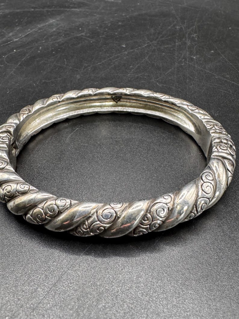 Puede incluir: Un brazalete plateado con un dise&ntilde;o de cuerda trenzada y adornos ornamentados. El brazalete tiene una marca en forma de coraz&oacute;n. El brazalete tiene aproximadamente 6,35 cm de di&aacute;metro, sobre un fondo oscuro.