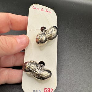 Può includere: Orecchini argentati a forma di arachide su una scheda bianca Casa de Leon. Gli orecchini presentano superfici strutturate. La scheda riporta il testo "Casa de Leon" e "U.S.A."