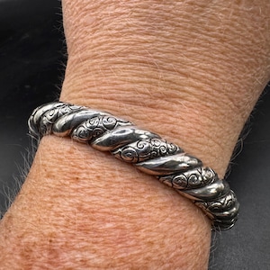 Puede incluir: Un brazalete plateado con un dise&ntilde;o de cuerda trenzada. El brazalete presenta intrincados adornos y se lleva en la mu&ntilde;eca. El metal tiene un acabado pulido, que refleja la luz. El brazalete es una pieza de joyer&iacute;a.