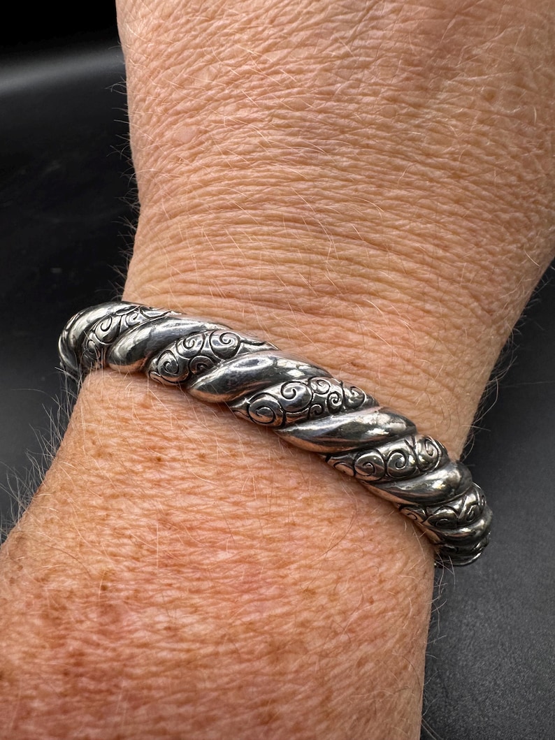 Puede incluir: Un brazalete plateado con un dise&ntilde;o de cuerda trenzada. El brazalete presenta intrincados adornos y se lleva en la mu&ntilde;eca. El metal tiene un acabado pulido, que refleja la luz. El brazalete es una pieza de joyer&iacute;a.