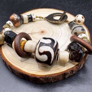 May include: A bracelet featuring a central large white bead with a dark brown swirling pattern, flanked by dark brown wooden rings, faceted black beads, and smaller white, metallic copper, and textured grey beads, secured with a toggle clasp.