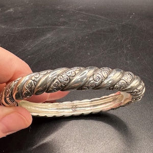 Puede incluir: Un brazalete plateado con un dise&ntilde;o de cuerda trenzada y adornos ornamentales. El brazalete tiene un cierre de bisagra y est&aacute; sostenido por una mano sobre un fondo oscuro. Es una pieza de joyer&iacute;a.