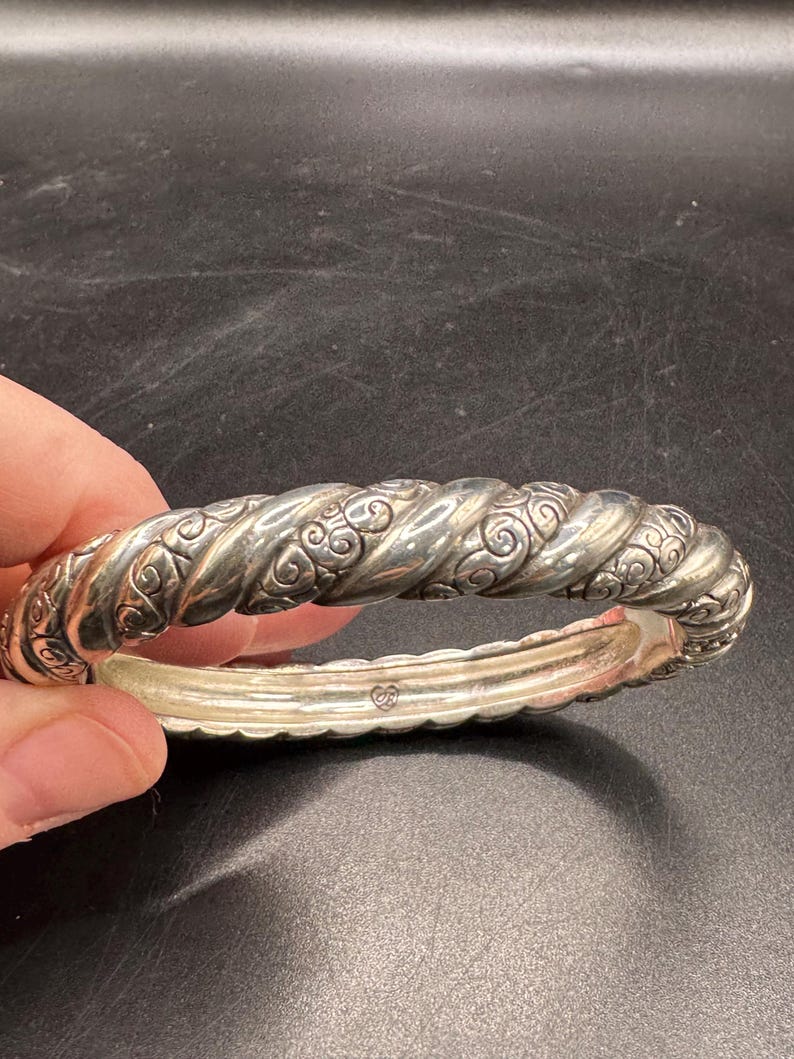Puede incluir: Un brazalete plateado con un dise&ntilde;o de cuerda trenzada y adornos ornamentales. El brazalete tiene un cierre de bisagra y est&aacute; sostenido por una mano sobre un fondo oscuro. Es una pieza de joyer&iacute;a.