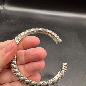 Puede incluir: Un brazalete plateado con un dise&ntilde;o de cuerda trenzada y adornos de volutas. El brazalete abierto se sostiene en una mano sobre un fondo oscuro. El brazalete es una pieza de joyer&iacute;a.