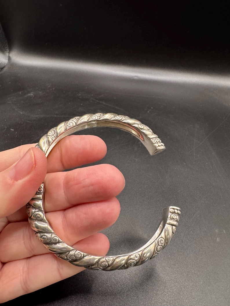 Puede incluir: Un brazalete plateado con un dise&ntilde;o de cuerda trenzada y adornos de volutas. El brazalete abierto se sostiene en una mano sobre un fondo oscuro. El brazalete es una pieza de joyer&iacute;a.
