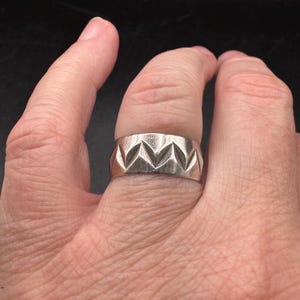 May include: A wide silver-toned metal band ring featuring a repeating pattern of recessed, triangular geometric carvings that create a series of V-shaped indentations around the entire circumference of the polished metallic surface.