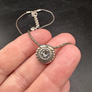 May include: Silver necklace with a round pendant. The pendant has a central clear stone and a decorative border. The chain is a fine, woven design. The clasp is visible. The necklace is displayed against a dark background.