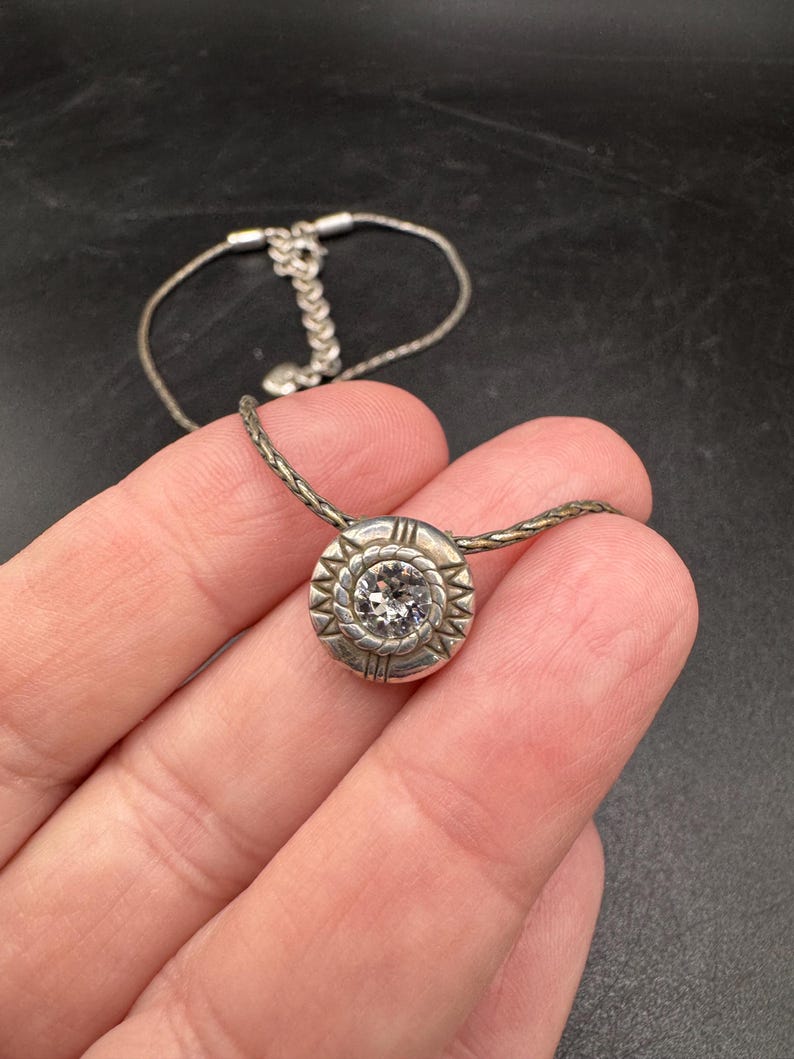 May include: Silver necklace with a round pendant. The pendant has a central clear stone and a decorative border. The chain is a fine, woven design. The clasp is visible. The necklace is displayed against a dark background.