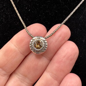 May include: A silver necklace with a circular pendant. The pendant has a central, round, light brown gemstone, surrounded by a decorative silver border with a starburst pattern. The chain is a fine, textured silver.