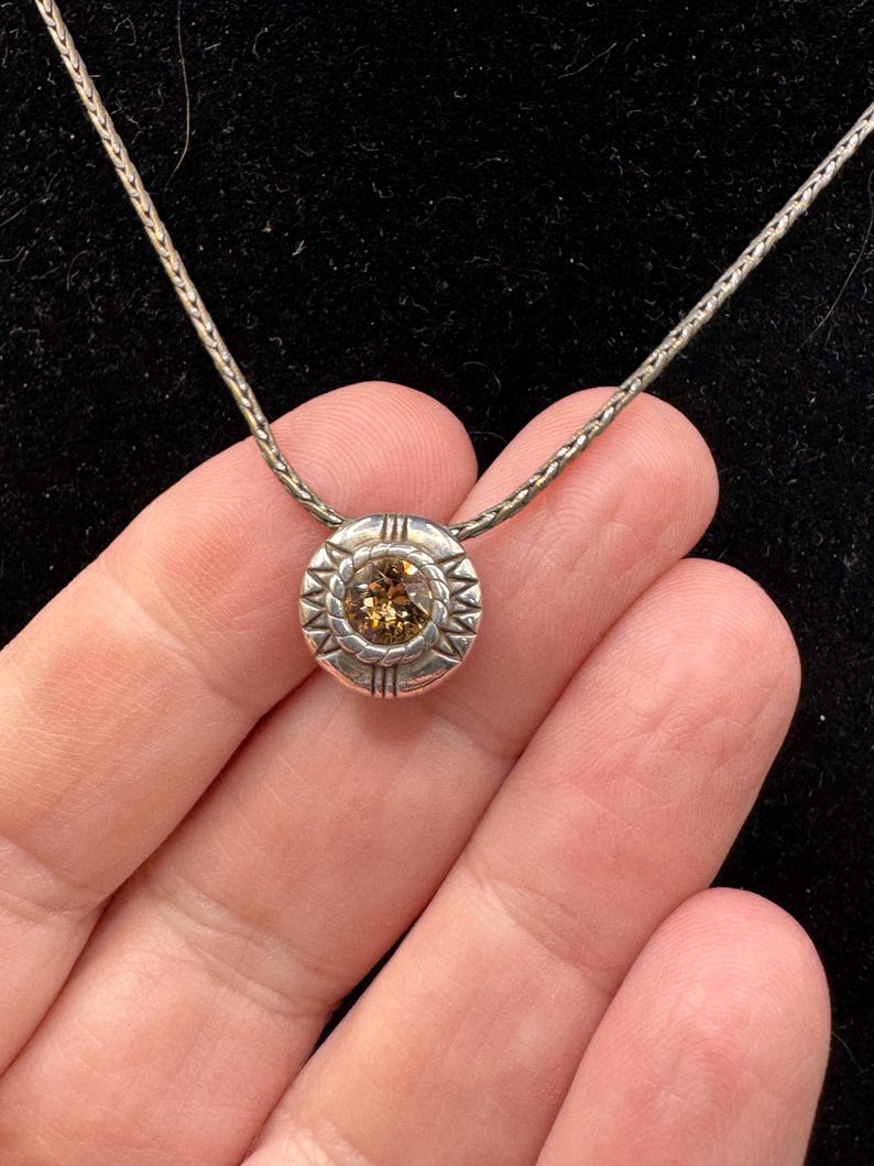 May include: A silver necklace with a circular pendant. The pendant has a central, round, light brown gemstone, surrounded by a decorative silver border with a starburst pattern. The chain is a fine, textured silver.