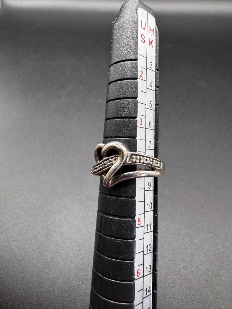 May include: A silver ring with a heart design and small clear stones. The ring is on a sizing tool and measures size 6.