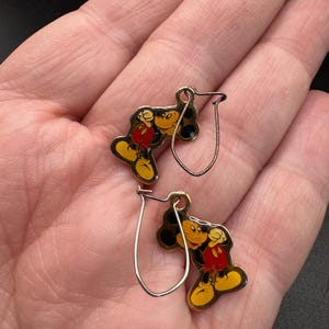 K&ouml;nnte beinhalten: Ein Paar Mickey Mouse-Ohrringe. Jeder Ohrring hat einen gelben, roten, schwarzen und wei&szlig;en Emaille-Mickey-Mouse-Anh&auml;nger mit einem silberfarbenen Haken. Die Ohrringe werden auf hellem Hintergrund pr&auml;sentiert.