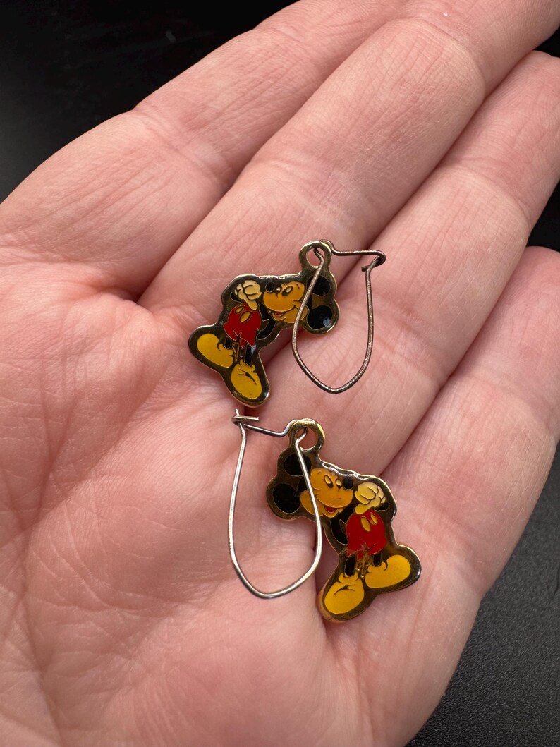 K&ouml;nnte beinhalten: Ein Paar Mickey Mouse-Ohrringe. Jeder Ohrring hat einen gelben, roten, schwarzen und wei&szlig;en Emaille-Mickey-Mouse-Anh&auml;nger mit einem silberfarbenen Haken. Die Ohrringe werden auf hellem Hintergrund pr&auml;sentiert.