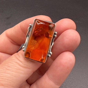 May include: A rectangular ring featuring a large, polished amber stone with deep orange and golden hues. The stone is set in a silver metal frame with decorative beaded accents along the sides, showcasing natural inclusions within the amber.