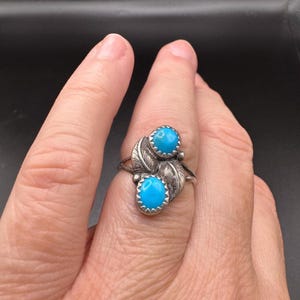 May include: A silver ring featuring two oval, bright turquoise stones. The stones are set in a decorative setting with leaf-like details. The ring is shown on a finger.