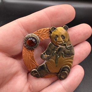 May include: A circular brooch featuring a metallic panda bear eating bamboo, set against a textured copper-toned background. The design includes a small, round, dark amber-colored cabochon stone embedded in the upper left side of the frame.