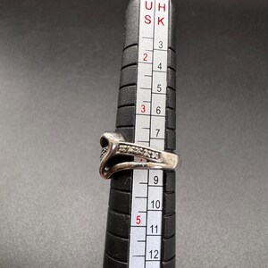 May include: A silver ring with a heart-shaped design and small clear stones. The ring is sized on a ring sizer that shows sizes 5 through 12.