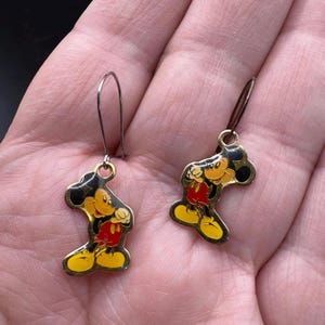 K&ouml;nnte beinhalten: Ein Paar Vintage-Ohrringe mit Mickey Mouse-Anh&auml;ngern. Jeder Ohrring hat eine goldfarbene Metallkontur von Mickey Mouse mit schwarzen, gelben, roten und orangefarbenen Emaille-Details. Die Ohrringe haben einen einfachen Hakenverschluss.