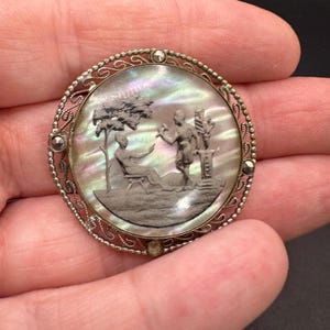 Op de afbeelding: Een ovale zilveren broche met een cameo van twee figuren in een tuinsetting. De cameo is gezet in een filigraan frame met een parelmoerachtige, iriserende achtergrond.