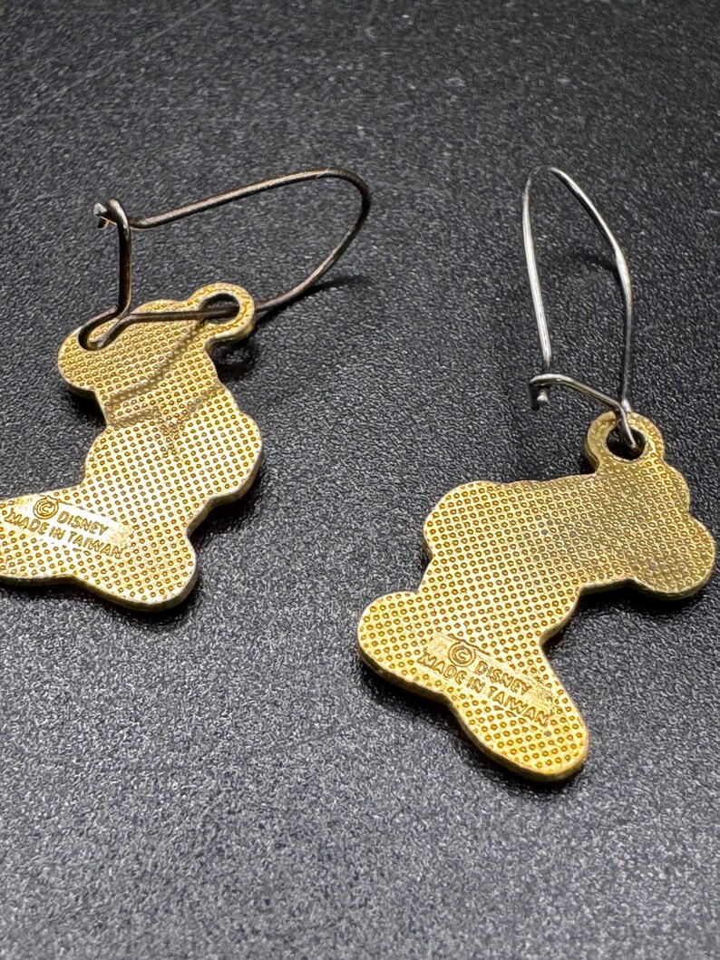 K&ouml;nnte beinhalten: Ein Paar goldfarbene Ohrh&auml;nger. Jeder Ohrring hat eine stilisierte Form mit strukturierter Oberfl&auml;che und einem Drahtb&uuml;gel. Die Ohrringe sind mit "Disney Made in Taiwan" gekennzeichnet.