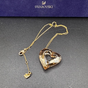 Peut inclure: Un collier à chaîne dorée avec un pendentif en cristal ambré à facettes en forme de cœur évidé. Le collier comprend un fermoir mousqueton et une petite breloque près de la fermeture gravée du logo cygne de Swarovski.