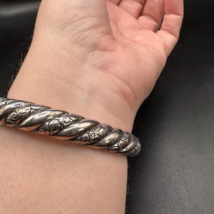 Puede incluir: Un brazalete plateado con un dise&ntilde;o de cuerda trenzada y detalles florales ornamentados. El brazalete se muestra en una mu&ntilde;eca, mostrando su textura y artesan&iacute;a. La pieza de joyer&iacute;a tiene una est&eacute;tica vintage.