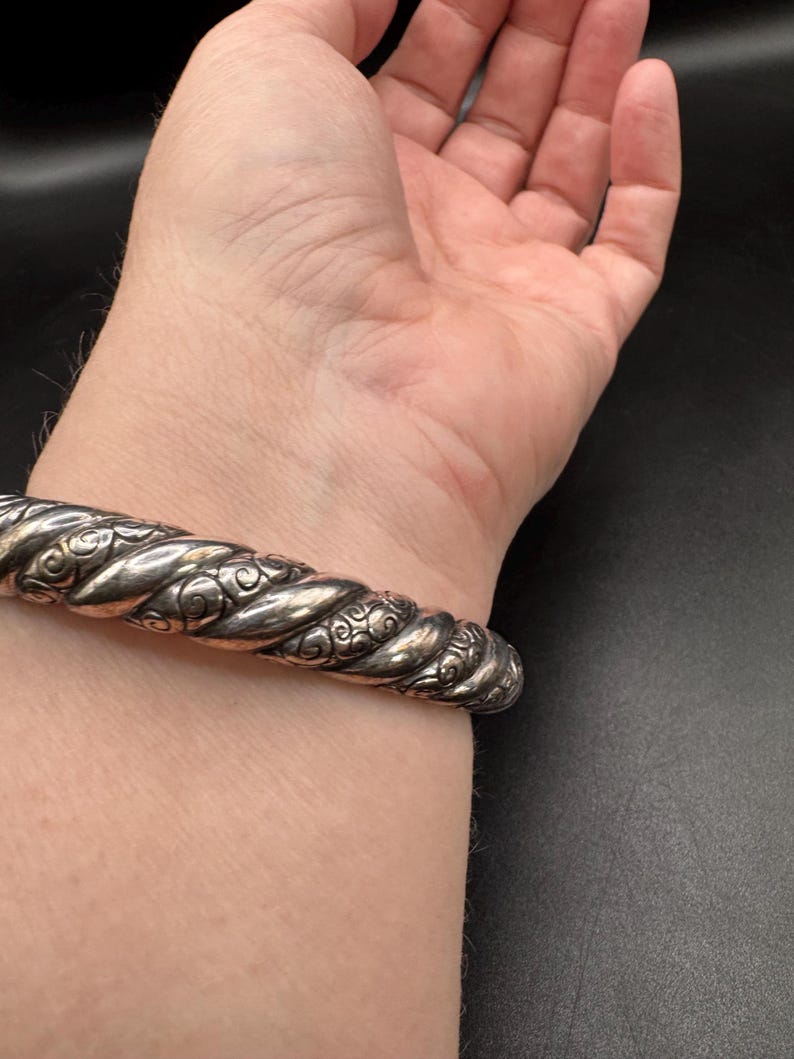 Puede incluir: Un brazalete plateado con un dise&ntilde;o de cuerda trenzada y detalles florales ornamentados. El brazalete se muestra en una mu&ntilde;eca, mostrando su textura y artesan&iacute;a. La pieza de joyer&iacute;a tiene una est&eacute;tica vintage.
