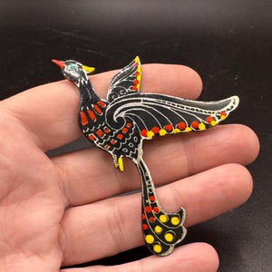 Può includere: Spilla decorativa a forma di uccello, con base nera e dettagli rossi, gialli e bianchi. L'uccello ha un becco rosso, un occhio verde e dettagli gialli sulla testa e sulle ali. La spilla è tenuta in mano su sfondo scuro.