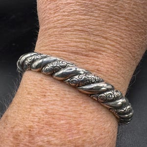 Puede incluir: Un brazalete plateado con un dise&ntilde;o de cuerda trenzada y adornos de volutas. El brazalete se lleva en la mu&ntilde;eca, mostrando su textura detallada y artesan&iacute;a. Una pieza de joyer&iacute;a cl&aacute;sica.