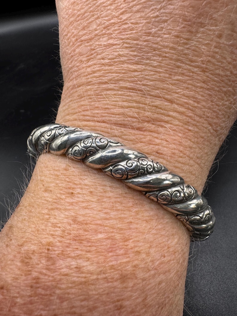 Puede incluir: Un brazalete plateado con un dise&ntilde;o de cuerda trenzada y adornos de volutas. El brazalete se lleva en la mu&ntilde;eca, mostrando su textura detallada y artesan&iacute;a. Una pieza de joyer&iacute;a cl&aacute;sica.