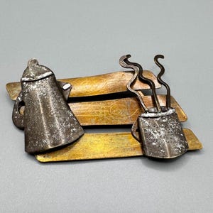 May include: A metal brooch depicting a coffee pot and a cup of coffee with steam rising from it. The coffee pot and cup are made of silver metal and are sitting on a bench made of gold metal.