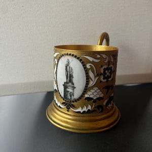 Peut inclure: Un porte-tasse en métal doré avec une poignée. Le porte-tasse présente une image en noir et blanc d'une statue dans un cadre ovale, entourée de motifs floraux blancs et noirs.